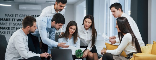 Guy shows document to a girl. Group of young freelancers in the office have conversation and working.